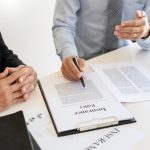 Two individuals discussing insurance claims documentation, with one person pointing at a contract on the table, emphasizing claims adjustment and representation.